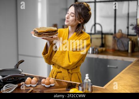 Portrait einer jungen fröhlichen Hausfrau in Bademantel mit einem Stapel gebratener Pfannkuchen gekleidet, das Frühstück in der Küche zu Hause vorbereiten. Kochen gesunde Lebensmittel und glücklich Hausarbeit Konzept Stockfoto