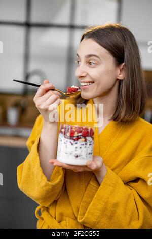 Junge Frau im gelben Bademantel genießt gesundes Müsli-Frühstück mit Joghurt und Beeren in der Schüssel in der Küche zu Hause am Morgen. Nachdenklicher lächelnder Look. Stockfoto