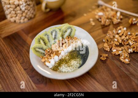 Gesundes Frühstück mit Müslischale mit Kiwi, Spirulina und Bio-Joghurt auf Holzplatte in der Küche mit Nüssen auf dem Hintergrund. Stockfoto