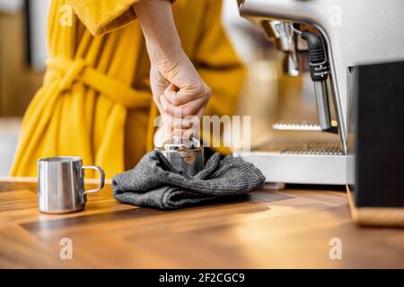 Frau im gelben Bademantel macht Espresso-Getränk auf einer professionellen Kaffeemaschine zu Hause, Nahaufnahme. Der Prozess der Herstellung von Kaffee auf einer Johannisbrotmaschine. Stockfoto