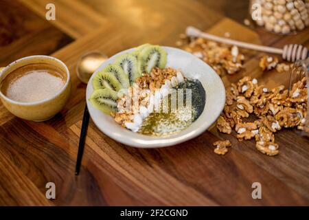 Gesundes Frühstück mit Müslischale mit Kiwi, Spirulina und Bio-Joghurt auf Holzplatte in der Küche mit Nüssen und Kaffee auf dem Hintergrund. Stockfoto