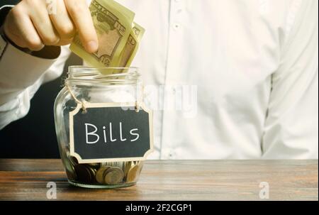 Ein Mann steckt Geld in ein Glas mit dem Wort Rechnungen. Zahlungskonzept für Stromrechnung. Gehäuse, Strom, Wasser Rechnungen. Konto, Geschäft und Finanzen. S Stockfoto