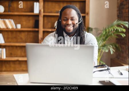 Vorderansicht eines afroamerikanischen Mannes, der einen Laptop und ein Freisprechgerät für die Online-Kommunikation mit Kollegen, Kunden, die am Schreibtisch im Büro sitzen, einen biracial lächelnden Kerl, der ein Headset in der Halle trägt Stockfoto