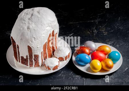 Hausgemachte handgemachte Osterkuchen mit weißer Glasur und bunt bemalten Ostereiern auf Tellern auf dunklem Hintergrund bedeckt. Stockfoto