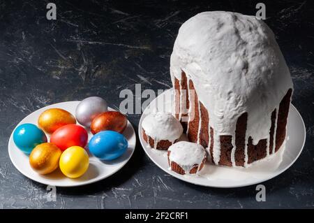 Hausgemachte handgemachte Osterkuchen mit weißer Glasur und bunt bemalten Ostereiern auf Tellern auf dunklem Hintergrund bedeckt. Stockfoto