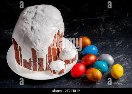Hausgemachte handgemachte Osterkuchen mit weißer Glasur und bunt bemalten Ostereiern auf Tellern auf dunklem Hintergrund bedeckt. Stockfoto