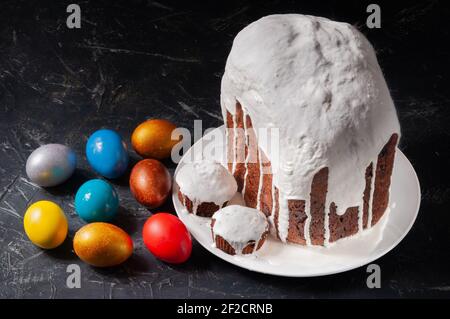 Hausgemachte handgemachte Osterkuchen mit weißer Glasur und bunt bemalten Ostereiern auf Tellern auf dunklem Hintergrund bedeckt. Stockfoto