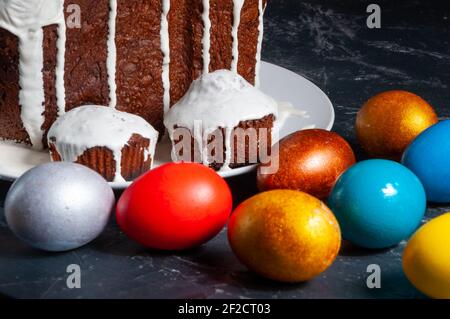 Hausgemachte handgemachte Osterkuchen mit weißer Glasur und bunt bemalten Ostereiern auf Tellern auf dunklem Hintergrund bedeckt. Stockfoto
