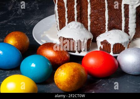 Hausgemachte handgemachte Osterkuchen mit weißer Glasur und bunt bemalten Ostereiern auf Tellern auf dunklem Hintergrund bedeckt. Stockfoto