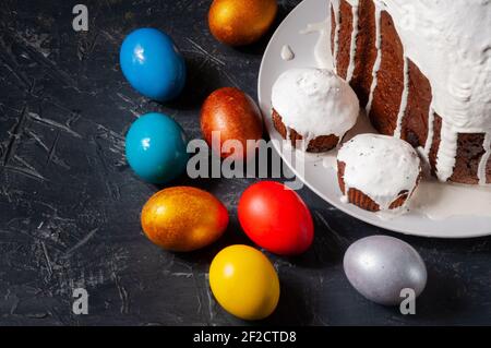 Hausgemachte handgemachte Osterkuchen mit weißer Glasur und bunt bemalten Ostereiern auf Tellern auf dunklem Hintergrund bedeckt. Stockfoto