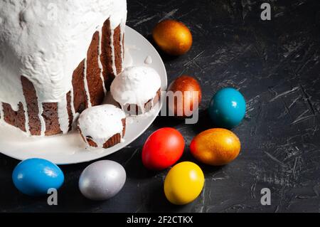 Hausgemachte handgemachte Osterkuchen mit weißer Glasur und bunt bemalten Ostereiern auf Tellern auf dunklem Hintergrund bedeckt. Stockfoto