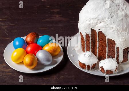 Hausgemachte handgemachte Osterkuchen mit weißer Glasur und bunt bemalten Ostereiern auf Tellern auf dunklem Holzhintergrund bedeckt. Stockfoto