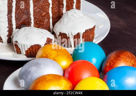 Hausgemachte handgemachte Osterkuchen mit weißer Glasur und bunt bemalten Ostereiern auf Tellern auf dunklem Holzhintergrund bedeckt. Stockfoto