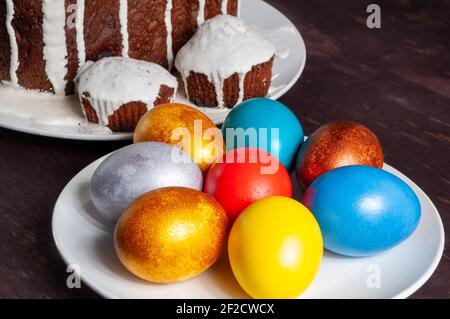 Hausgemachte handgemachte Osterkuchen mit weißer Glasur und bunt bemalten Ostereiern auf Tellern auf dunklem Holzhintergrund bedeckt. Stockfoto