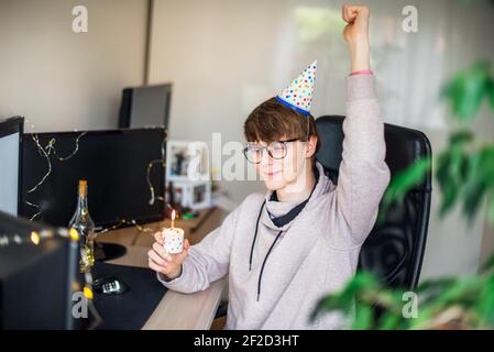 Emotionaler Mann in witziger B-Day Mütze mit festlichem Cupcake mit brennender Kerze auf virtueller Geburtstagsparty beim Lockdown. Generation Z Mann feiert Stockfoto