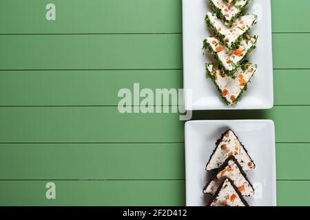 Schöne Snacks von weichem Frischkäse auf einem weißen Teller auf grünem Hintergrund. Hüttenkäse-Snacks mit Lachs und Kräutern. Stockfoto