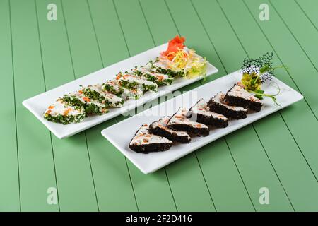 Schöne Snacks von weichem Frischkäse auf einem weißen Teller auf grünem Hintergrund. Hüttenkäse-Snacks mit Lachs und Kräutern. Stockfoto