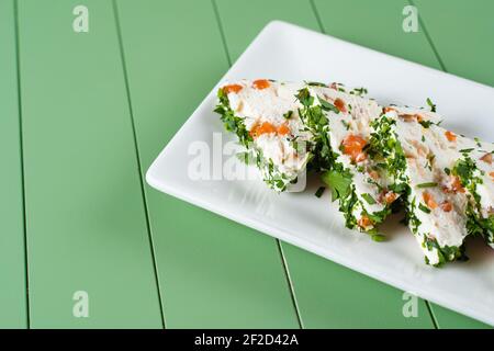 Schöne Snacks von weichem Frischkäse auf einem weißen Teller auf grünem Hintergrund. Hüttenkäse-Snacks mit Lachs und Kräutern. Stockfoto