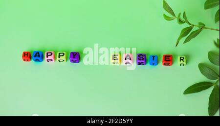 Frohe Ostern Worte geschrieben mit bunten Holz Alphabet Buchstaben in zufälliger Reihenfolge auf grünem Hintergrund flach. Grüner Zweig mit jungen Blättern. Natürliches E Stockfoto