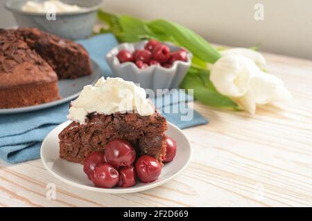 Saftige hausgemachte Schokoladenkuchen mit Morello-Kirschen und Schlagsahne, einige Tulpenblumen und eine blaue Serviette auf einem hellen Holztisch für einen festlichen Ader Stockfoto