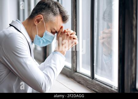 Frustriert erschöpft männlichen Arzt in einer medizinischen Uniform und schützende medizinische Maske. Ein müder Arzt steht am Fenster nach einem harten Arbeitstag, Überstunden, er hat Kopfschmerzen, braucht Ruhe Stockfoto