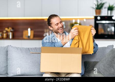 Lächelnder attraktiver Mann packte sein Paket aus und freute sich, eine lange erwartete Bestellung zu bekommen. Kaukasischen modernen Kerl Einkaufen in Internet-Shops, Kauf neuer Kleidung online, Online-Shopping-Konzept Stockfoto