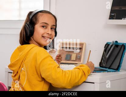 Kleine kaukasische Mädchen, mit einem Guss in einer Hand, trägt ein gelbes Sweatshirt, Skizzen an ihrem Schreibtisch sehr glücklich und lächelnd, mit Zeichenmaterial Stockfoto