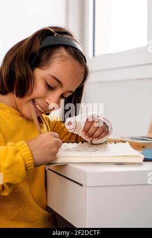 Kleine kaukasische Mädchen, mit einem Guss in einer Hand, trägt ein gelbes Sweatshirt, Skizzen an ihrem Schreibtisch sehr glücklich und lächelnd, mit Zeichenmaterial Stockfoto