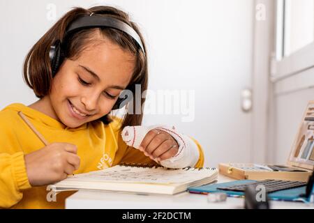 Kleine kaukasische Mädchen, mit einem Guss in einer Hand, trägt ein gelbes Sweatshirt, Skizzen an ihrem Schreibtisch sehr glücklich und lächelnd, mit Zeichenmaterial Stockfoto