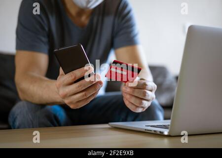 Glatze Mann in einer medizinischen Maske mit einer Kreditkarte sitzt auf der Couch an einem Laptop mit einem Telefon in der Hand, das Konzept des Online-Shopping in einem Online-Shop Stockfoto