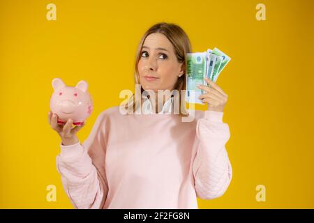 Porträt einer nachdenklichen jungen Frau hält Sparschwein und Bündel von Geldscheinen isoliert über gelben Hintergrund Stockfoto
