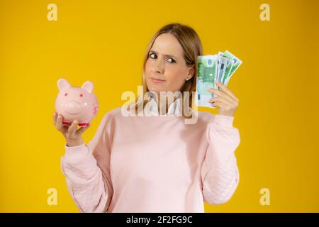 Porträt einer nachdenklichen jungen Frau hält Sparschwein und Bündel von Geldscheinen isoliert über gelben Hintergrund Stockfoto
