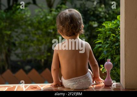 Ein charmantes Kleinkind sitzt mit einem Spielzeug auf den Stufen eines Hauses im Sonnenlicht. Rückansicht. Stockfoto