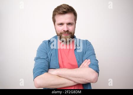 Ein attraktiver Kaukasier mit wohlwollender Erscheinung steht mit gekreuzten Armen und schaut in die Kamera. Weißer Hintergrund. Stockfoto