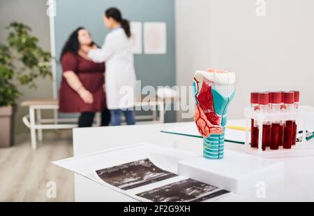Schilddrüsenanatomisches Modell und Schilddrüsenhormon-Blutteströhrchen, Nahaufnahme. Der Arzt untersucht den Hals einer fetten Frau im Hintergrund Stockfoto