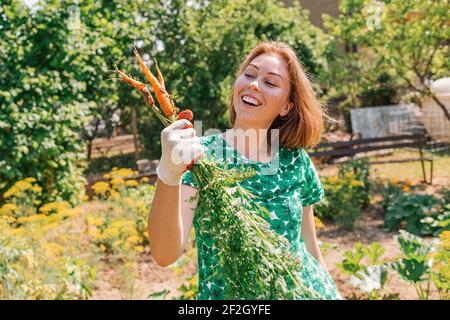 Eine kaukasische junge Frau hält freudig ein Bündel von Karotten aus dem Garten gesammelt.Vegetation im Hintergrund.Konzept der Ernte und Gartenarbeit. Stockfoto