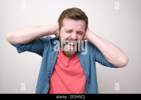 Kaukasischer Mann in Stress, er ist des Lärms müde, bedeckt Ohren mit Händen und schreit laut. Psychologisches Gesundheitskonzept. Stockfoto