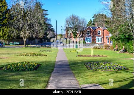 Ein Fußweg führt zur Boltro Road durch den Muster Green, einem kleinen Park in Haywards Heath, West Sussex, mit Leuten auf Bänken, die den Frühlingssonne genießen. Stockfoto