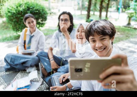 Ein gutaussehender männlicher Gymnasiast lächelt und hält ein Handy Während Sie ein Selfie mit einer Gruppe von Freunden machen Stockfoto