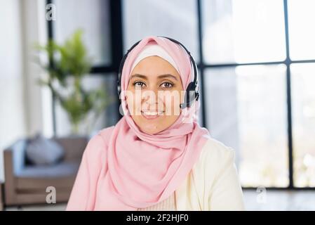 Kopfaufnahme einer jungen muslimischen arabischen Mischrassen-Frau in Hijab und Headset, die im Büro mit Panoramablick sitzt, in der Kundendienstabteilung als Callcenter-Betreiber arbeitet und die Kamera anschaut Stockfoto