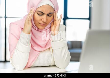 Müde muslimische Araber gemischt-Rasse weibliche Angestellte Geschäftsfrau in Hijab schloss ihre Augen berühren massierenden Tempeln, leiden unter schweren Kopfschmerzen Migräne, Gefühl überwältigt von der Arbeit auf Laptop Stockfoto