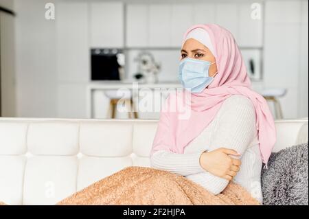 Junge muslimische arabische Frau in Hijab und schützende Gesichtsmaske sitzen auf Couch halten sich, die Beine mit Decke, Gefühl kalt krank von Grippe, Selbstisolation und Coronavirus-Behandlungskonzept Stockfoto