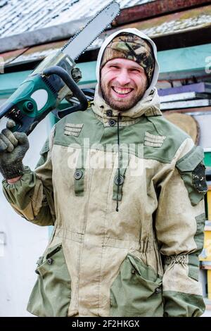 Attraktiver Mann in Camouflage mit Kettensäge und lächelt. Bärtiger Mann, der im Winter in der Nähe der Scheune auf dem Land steht Stockfoto