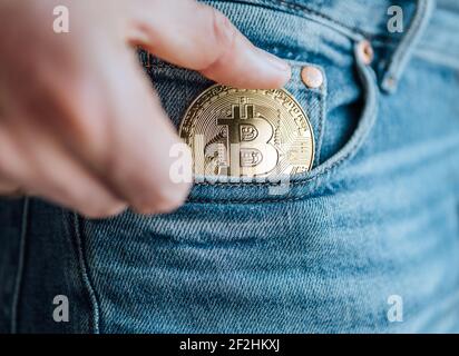 Nahaufnahme von Männerfingern, die die Bitcoin-Münze in die blaue Jeans-kleine Tasche stecken. Ersparnisse, digitale Währung, Kryptowährung, Investitionen und s Stockfoto