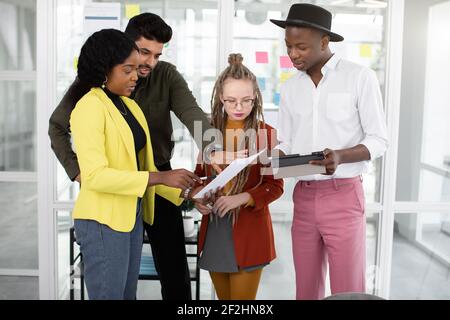 Vier multiethnische kreative junge Kollegen in lässigen, stylischen Outfits diskutieren über ihr neues Startup, schauen sich Papier und Tablet an Stockfoto