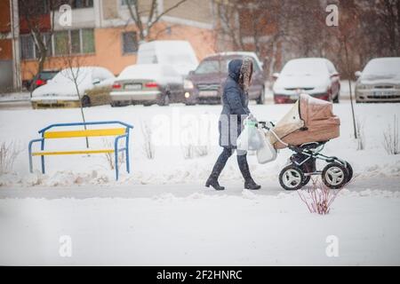 Eine Frau mit Kinderwagen läuft auf einer verschneiten Straße. Wandern mit Kind bei jedem Wetter. Mit einem Kinderwagen durch die Schneeverwehungen. Zu Fuß Neugeborene in der Stockfoto