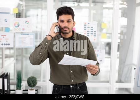 Junger gutaussehender indischer Geschäftsmann telefonieren, posiert im modernen Büro mit Papieren Stockfoto