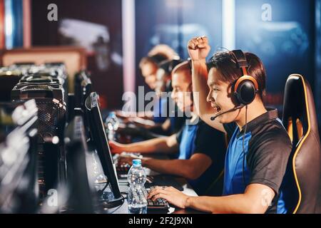 Seitenansicht der aufgeregt jungen asiatischen Kerl, männliche Cyber-Sport-Gamer Blick auf PC-Bildschirm und Anheben Hand nach oben, während Stockfoto