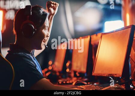 Seitenansicht des jungen aufgeregt asiatischen Kerl, männlich professionellen Cybersport Gamer trägt Kopfhörer heben Hand nach oben, wining in eSport-Turnier Stockfoto