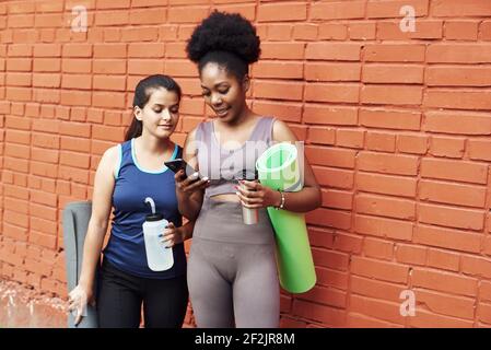 Bild von erstaunlichen jungen athletischen Frauen vor einer Ziegelwand, Blick in das Telefon. Zwei Freundinnen lachen über den Social Media Post. Stockfoto